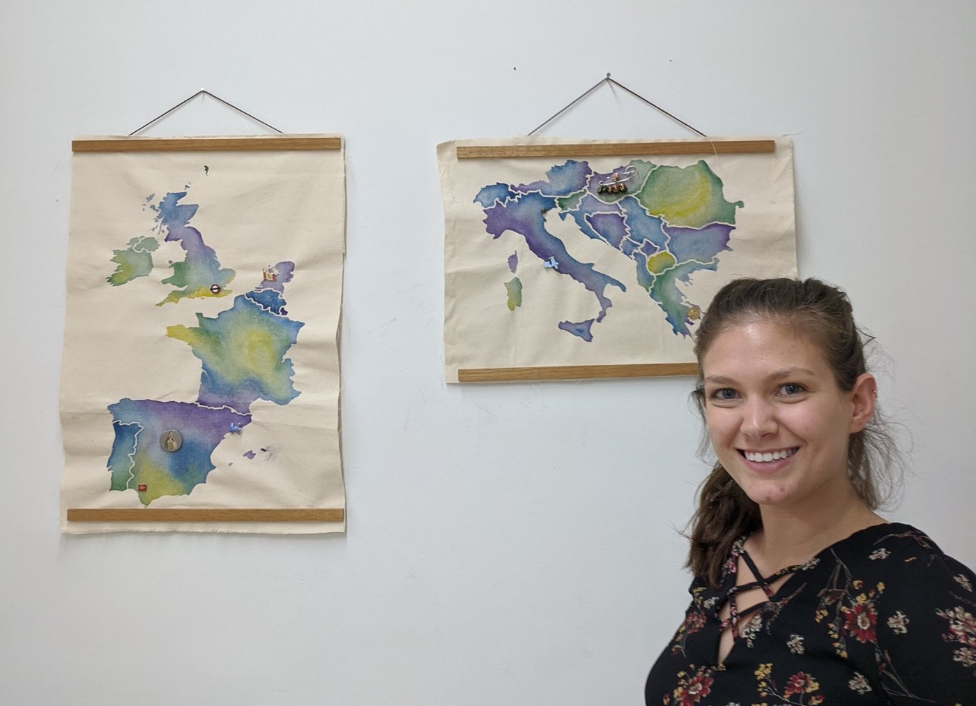 Laura Bloch standing beside two finished watercolor canvas maps hanging on a wall — western Europe and central Europe